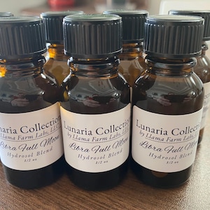 May include: A collection of small amber glass bottles with black caps. Each bottle has a white label with the text "Lunaria Collection Libra Full Moon Hydrosol Blend 1/2 oz". The bottles are arranged on a light brown surface.