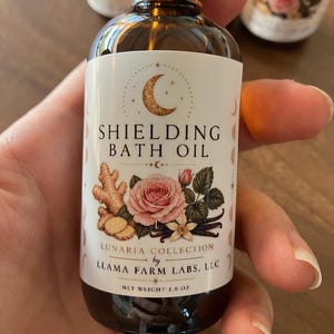 May include: A brown glass bottle of Shielding Bath Oil with a black cap. The label features a crescent moon, a pink rose, ginger, and vanilla. The net weight is 1.8 oz.
