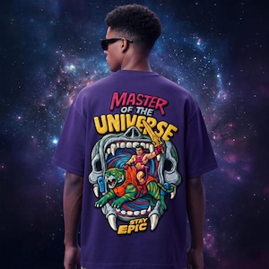 May include: A purple t-shirt featuring a graphic from the Masters of the Universe franchise. The design includes the text "MASTER OF THE UNIVERSE" and an illustration of a character riding a tiger. The phrase "STAY EPIC" is also present.