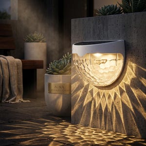 May include: A white solar-powered outdoor wall light with a textured, geometric design. The light is mounted on a concrete wall, casting a warm glow and patterned shadows. The light is labeled "AURA GEO-FACETED LIGHT".