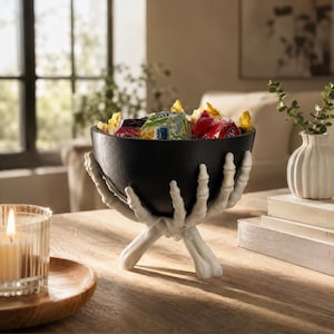 May include: A black bowl held by a white skeletal hand base, filled with assorted colourful wrapped sweets. The bowl has a smooth, matte finish, while the base features detailed finger bones and knuckles forming a sturdy stand for the container.