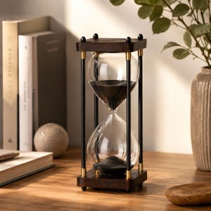 Könnte beinhalten: Eine Sanduhr mit einem klaren Glaskörper gefüllt mit schwarzem Sand, eingefasst in einen Rahmen aus dunklem Holz mit quadratischen Platten oben und unten, verbunden durch vier schlanke schwarze Metallstäbe mit goldfarbenen Akzenten.