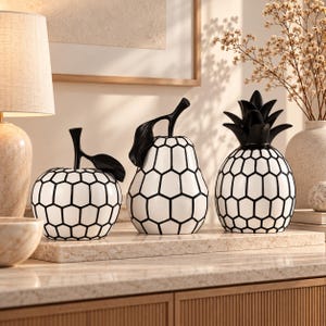 May include: Three decorative fruit sculptures featuring a white base with a black honeycomb pattern. The set includes an apple, a pear, and a pineapple, each topped with a solid black stem or leaves, arranged in a row on a flat rectangular surface.