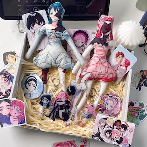 May include: A white gift box filled with shredded paper containing two fabric dolls, one in a blue dress and one in a pink dress, surrounded by various anime-style character stickers, circular pin-back buttons, and acrylic keychains featuring colorful illustrations.