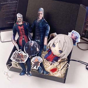 May include: A collection of merchandise featuring two flat fabric dolls of male characters with silver hair, a round button pin, a plush doll with white hair, and several small stickers, all arranged inside a black cardboard gift box.