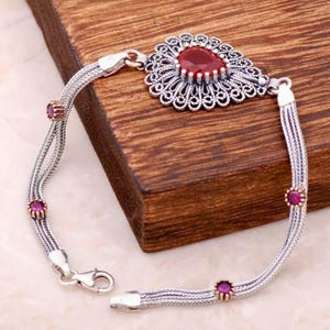May include: A silver bracelet featuring a central teardrop-shaped red stone surrounded by intricate filigree metalwork. The chain consists of three braided silver strands accented with four small, round, bezel-set red stones along the length.