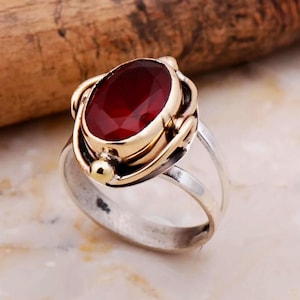 Peut inclure: Bague en argent avec une grosse pierre précieuse rouge foncé de forme ovale, sertie dans une lunette dorée. La bague présente un design à tige fendue et des accents dorés. La pierre précieuse à facettes est le point central.