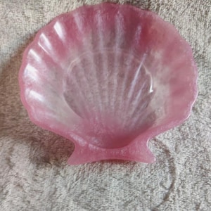 Può includere: Un piatto rosa a forma di conchiglia con finitura perlata. Il piatto ha un bordo smerlato e una superficie strutturata, che ricorda una vera conchiglia. Il piatto è realizzato in un materiale solido e ha una piccola base piatta.