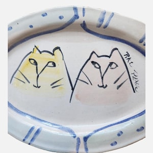 Op de afbeelding: Ovale keramische plaat met een handgeschilderd ontwerp met twee gestileerde kattengezichten. De katten zijn geel en roze, met zwarte contouren. De plaat heeft een blauwe rand met een gestippeld en gestreept patroon.