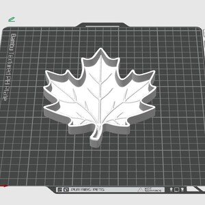 Puede incluir: Un cortador de galletas con forma de hoja de arce impreso en 3D en color blanco, centrado sobre una placa de construcci&oacute;n gris oscuro con patr&oacute;n de rejilla. La placa muestra el texto Bambu Textured PEI Plate y etiquetas de material.