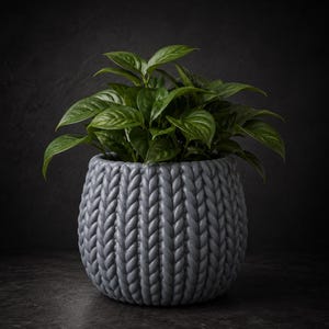 May include: A gray, textured, round planter pot with a knitted design, filled with vibrant green leafy plants. The pot is made of a durable material and is suitable for indoor or outdoor use. The background is a dark gray.