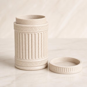 May include: A cylindrical beige canister with a speckled finish, featuring vertical fluted columns and a Greek key pattern border at the top and bottom. The canister has a threaded opening and a matching circular lid placed beside it on the surface.