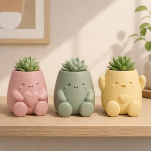 May include: Three small, rounded planters in pastel pink, sage green, and soft yellow, each featuring a sculpted character with a smiling face, arms, and legs. Each planter holds a green succulent plant and displays a heart detail on the front.