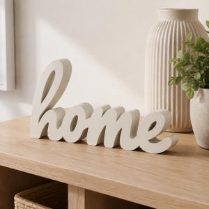 May include: A freestanding, off-white decorative sign featuring the word home in a lowercase, cursive script font. The letters are connected in a single, solid piece with a smooth, matte finish, designed to sit upright on a flat surface.