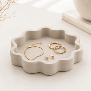 Op de afbeelding: Een lichtgrijze, ronde schaal met een geschulpte rand die een collectie goudkleurige sieraden bevat, waaronder een fijne kettingarmband, twee eenvoudige ringen en een paar kleine oorknopjes met heldere, fonkelende steentjes.