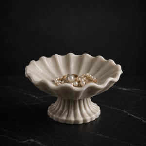 May include: A cream-colored, scalloped-edge pedestal bowl made of smooth stone, featuring a fluted design on both the basin and the base. Inside the bowl rests a gold ring with a central pearl and a strand of white pearls.