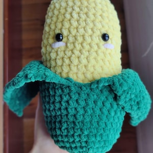 Könnte beinhalten: Ein handgehäkeltes Mais-Plüschtier mit einem lächelnden Gesicht. Das Plüschtier hat einen gelben Maiskornkörper und eine grüne Hülle. Die Augen sind schwarz mit weißen Akzenten. Das Plüschtier ist aus weichem, strukturiertem Garn gefertigt.