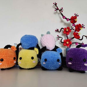 May include: A collection of five colorful, handmade crochet plush toys in orange, yellow, blue, pink, and purple. Each has black arms and legs, and a cute face. A decorative branch with red flowers is in the background.