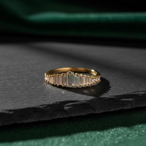 Peut inclure: Une bague en or ornée d'une rangée de pierres précieuses claires de coupe rectangulaire et ronde. La bague est posée sur une surface gris foncé avec un fond vert. Les pierres sont disposées symétriquement.