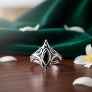 Peut inclure: Une bague en argent avec un design orné et une pierre noire. La bague a un sommet pointu et une bande texturée détaillée. Le fond comprend un tissu vert foncé et des pétales de fleurs blanches.