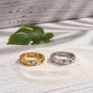 May include: Two rings, one gold and one silver, are displayed on a white surface. The gold ring features a teardrop-shaped pink stone, a small green stone, and blue accents. The silver ring has a similar design with a pink teardrop stone and blue accents.
