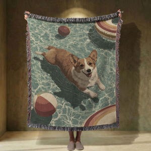 May include: A woven throw blanket featuring a corgi dog swimming in a pool with beach balls and a rainbow float. The blanket has a teal background with a fringed edge.