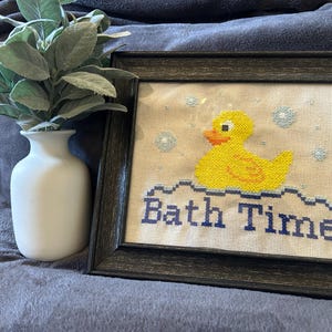 Puede incluir: Arte de punto de cruz enmarcado con un patito de goma amarillo sobre olas azules con las palabras "Bath Time" en azul marino. La obra de arte está en un marco gris oscuro, junto a un jarrón blanco con follaje verde.