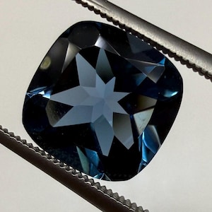 Könnte beinhalten: Ein facettierter, kissenförmiger Edelstein in tiefblauer Farbe, gehalten von einer Pinzette. Das Innere des Edelsteins weist ein sternförmiges Muster auf, das das Licht reflektiert. Die Pinzette ist silberfarben.