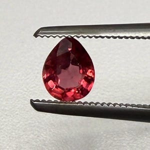 May include: A pear-shaped, faceted gemstone in a vibrant red hue is held by a pair of tweezers. The gem's polished surface reflects light, showcasing its clarity and brilliance. The tweezers are silver-colored and have a serrated edge.