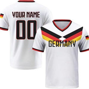 May include: A white V-neck jersey featuring a black, red, and yellow chevron pattern across the chest with the word GERMANY printed below. The back displays the text YOUR NAME and the number 00 in black with red outlines, plus a small German flag.