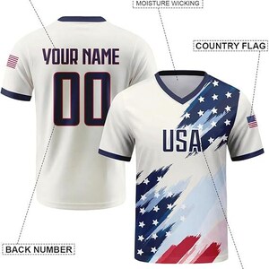 May include: A white V-neck jersey featuring a blue and red brushstroke American flag pattern on the front with the text USA. The back displays the text YOUR NAME and the number 00. Labels indicate breathable moisture wicking fabric features.