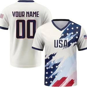 May include: A short-sleeved V-neck jersey featuring a cream base with a blue and red brushstroke flag pattern, the text USA, and a small flag on the sleeve. The back displays the text YOUR NAME and the number 00 in dark blue with red outlines.