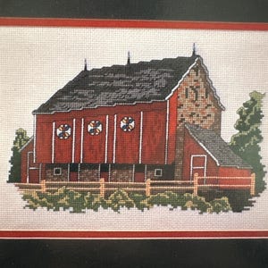 Puede incluir: Obra de arte de punto de cruz de un granero rojo con techo gris, tres ventanas redondas y un pequeño edificio adjunto. El granero está rodeado por una valla de madera y follaje verde. La obra está enmarcada en negro.