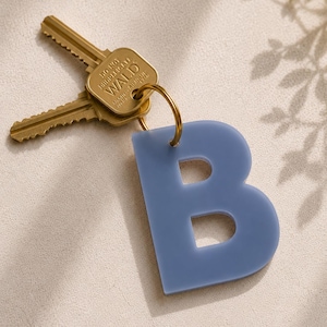 May include: A dusty blue acrylic letter B keychain attached to a gold-toned metal ring with two brass-colored keys. The key head is embossed with the text WALD and DO NOT DUPLICATE. The keychain features a smooth, matte finish and a simple design.