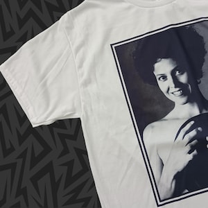 May include: White t-shirt featuring a black and white portrait of a woman. The portrait is framed with a black border. The woman has dark hair and is smiling. The t-shirt is made of a soft material.
