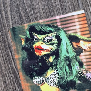 May include: A square sticker featuring a Gremlin character with long green hair, bright red lipstick, and white eyeshadow. The character wears a black and white leopard print bikini top. The background shows a blurred horizontal slat window blind.