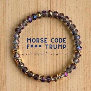 May include: A beaded bracelet with iridescent purple and clear beads, interspersed with gold-colored spacers. The bracelet features the text "MORSE CODE F*** TRUMP" in blue, along with the corresponding Morse code symbols.
