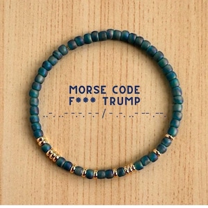 May include: A teal beaded bracelet with gold accents. The bracelet spells out "MORSE CODE F*** TRUMP" in Morse code. The bracelet is on a light brown wooden surface.