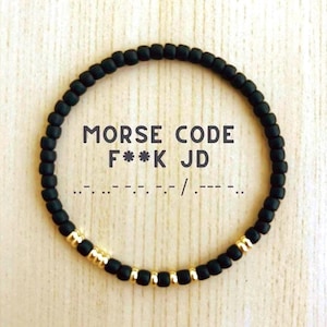 May include: A circular bracelet made of small matte black beads interspersed with metallic gold beads. The text MORSE CODE F**K JD is printed in the center, accompanied by the corresponding Morse code sequence in black dots and dashes.