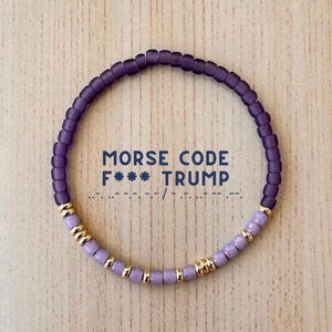 May include: A beaded bracelet with the text "MORSE CODE F*** TRUMP". The bracelet features purple and lavender beads with gold accents. The beads are strung together to form a circular shape.