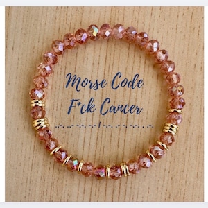 May include: A circular bracelet made of faceted, translucent rose-coloured beads interspersed with small, polished gold-toned metal rings. The centre of the image features the text Morse Code F*ck Cancer in dark blue script above a Morse code sequence.