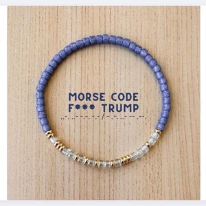 May include: A circular beaded bracelet featuring matte periwinkle blue beads along the top half and a sequence of clear glass and metallic gold spacer beads along the bottom half. The text MORSE CODE F*** TRUMP is printed in the centre.