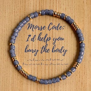 May include: A beaded bracelet with blue and gold beads. The bracelet has the text "Morse Code: I'd help you bury the body" in the center. Morse code symbols are below the text. The bracelet is on a wooden surface.