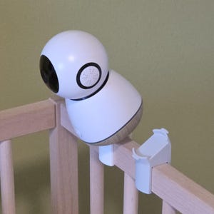 Könnte beinhalten: Ein weißer Babyphone-Monitor mit abgerundeter Oberseite und schwarzer Linse, befestigt an einem Holzbett. Der Monitor hat einen goldfarbenen Sockel und einen weißen Clip zur Befestigung.
