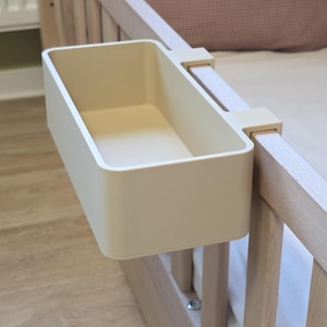 May include: A rectangular, beige plastic storage bin with a hook attachment designed to hang securely over the top rail of a wooden crib. The container features smooth, rounded corners and a deep, open-top design for holding small items.