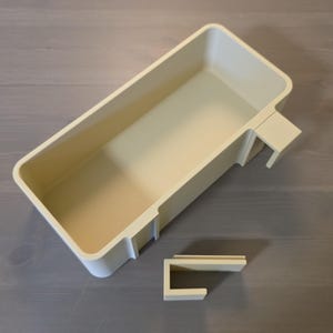 May include: A rectangular, off-white plastic storage bin featuring a deep, open interior and a hook-like attachment on one side. A separate, smaller, U-shaped plastic component in the same off-white color is positioned below the main bin.
