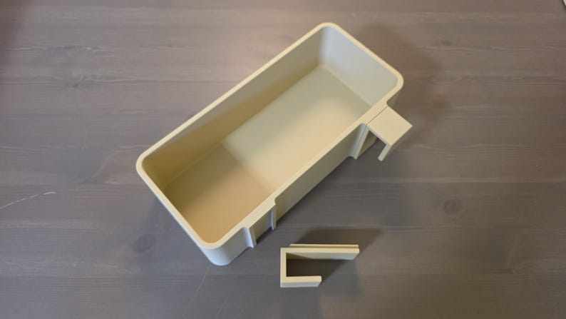 May include: A rectangular, off-white plastic storage bin featuring a deep, open interior and a hook-like attachment on one side. A separate, smaller, U-shaped plastic component in the same off-white color is positioned below the main bin.