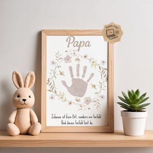 Könnte beinhalten: Ein gerahmter Druck mit dem Wort "Papa" und einem Handabdruck in der Mitte, umgeben von einem floralen Kranz. Der Druck enthält auch deutschen Text. Ein Plüschhase und eine Topf-Sukkulente stehen in der Nähe.