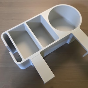 May include: A matte white desk organizer featuring a circular cup holder, two rectangular storage compartments, and a narrow vertical slot. The organizer includes an integrated clamp-style attachment arm designed for mounting onto a flat surface edge.