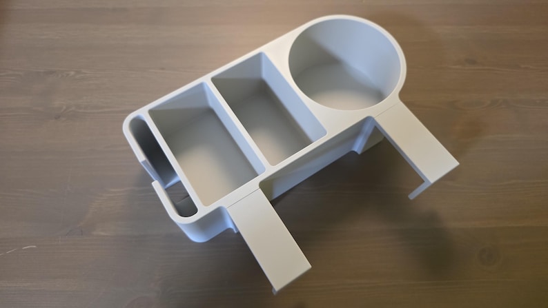 May include: A matte white desk organizer featuring a circular cup holder, two rectangular storage compartments, and a narrow vertical slot. The organizer includes an integrated clamp-style attachment arm designed for mounting onto a flat surface edge.